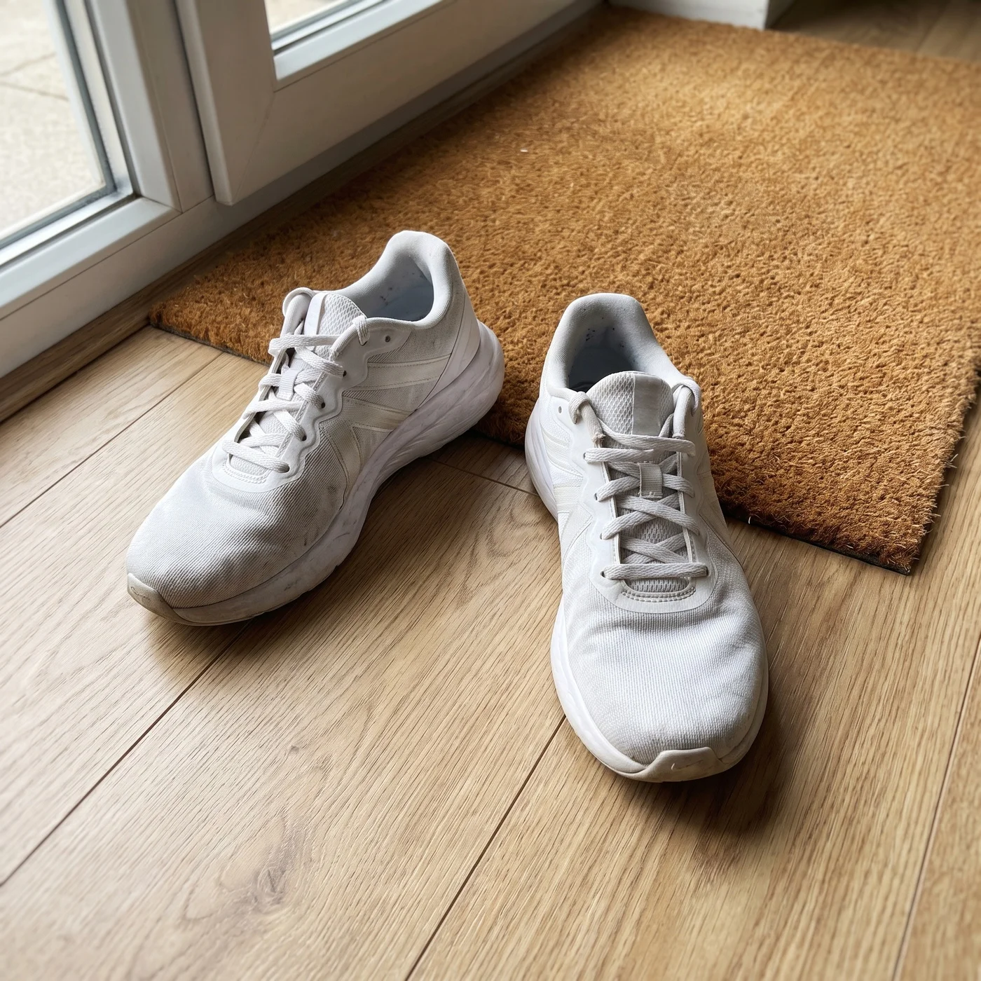 A pair of white running sneakers photographed on a wooden floor next to a doormat, casual seller snapshot in ambient home light